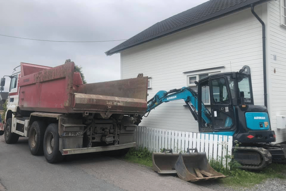 Lastebil og gravemaskin foran et hvitt hus